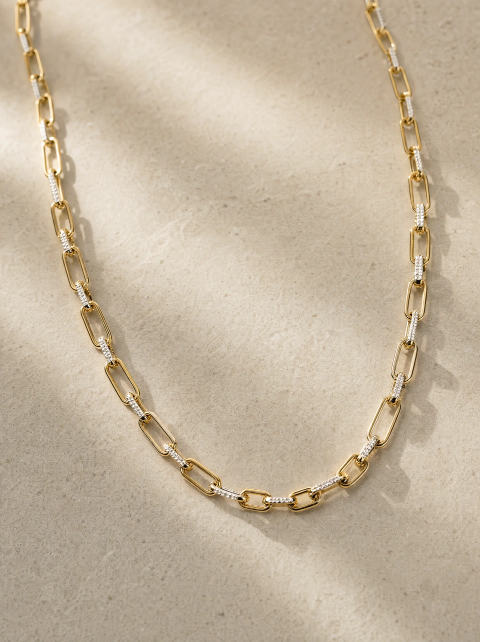 The paperclip necklace with gold-tone links and reflective worked-metal details, on a warm beige background